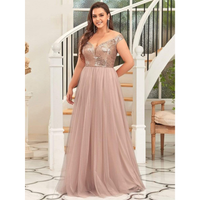 A woman in an elegant, off-the-shoulder dress with a sequin bodice and flowing blush skirt stands smiling in front of a white spiral staircase.