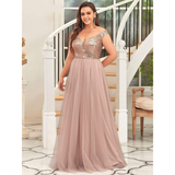 A woman in an elegant, off-the-shoulder dress with a sequin bodice and flowing blush skirt stands smiling in front of a white spiral staircase.