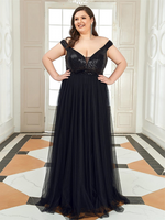 A woman in an elegant black off-the-shoulder gown with a sequined bodice stands confidently in a bright room, exuding joy and elegance.