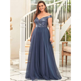 Woman in an elegant, off-the-shoulder blue gown with sequined bodice stands before a curved staircase, conveying a confident and joyful tone.