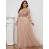 A woman in an off-the-shoulder, blush pink gown with a sequined bodice stands gracefully near a white fireplace. Her expression is warm and confident.