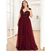 A woman in a burgundy evening gown with sequin details stands in a well-lit room with white walls and wooden floors, exuding elegance and confidence.