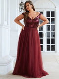 A woman in a burgundy gown with a sequined top and tulle skirt stands confidently in front of an elegant white building, conveying elegance and poise.