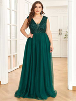 A woman poses confidently in an elegant, deep green evening gown with a sparkly bodice. The setting is a bright room with hardwood floors, conveying a formal and sophisticated mood.