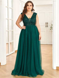 A woman poses confidently in an elegant, deep green evening gown with a sparkly bodice. The setting is a bright room with hardwood floors, conveying a formal and sophisticated mood.