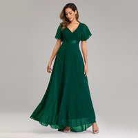 A woman in a flowing emerald green gown with flutter sleeves and a v-neckline stands gracefully. She wears silver sandals against a plain gray background.