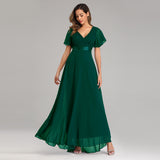 A woman in a flowing emerald green gown with flutter sleeves and a v-neckline stands gracefully. She wears silver sandals against a plain gray background.