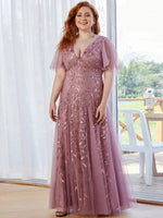 A woman in a mauve, floral-patterned gown stands in a bright room with sheer curtains. She smiles confidently, exuding elegance and warmth.