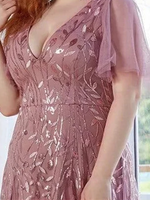 A woman in a mauve, leaf-patterned, sequin dress with sheer sleeves stands near a window. The dress conveys elegance and soft femininity.