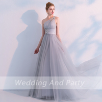 Elegant woman in a flowing, one-shoulder, light gray evening gown with jeweled details. She stands gracefully against a soft, pastel backdrop. 