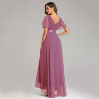 A woman in a flowing mauve gown with short flutter sleeves stands gracefully against a plain backdrop, exuding elegance and sophistication.