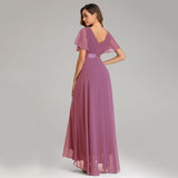 A woman in a flowing mauve gown with short flutter sleeves stands gracefully against a plain backdrop, exuding elegance and sophistication.