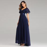 Woman in a floor-length, navy blue gown with flutter sleeves and a V-neckline, standing against a light gray background. She has a confident, elegant pose.