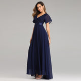 Woman in a floor-length, navy blue gown with flutter sleeves and a V-neckline, standing against a light gray background. She has a confident, elegant pose.