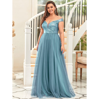 Woman in an elegant off-shoulder blue gown stands smiling beside a white staircase, exuding a formal and joyful atmosphere.