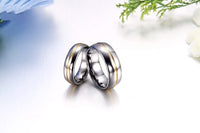 Two silver and gold wedding bands are intertwined on a reflective surface, with soft focus greenery and flowers in the background, conveying elegance and unity.