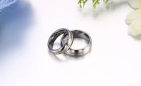 Two silver wedding bands lie intertwined on a smooth, reflective surface. A soft focus on white flowers and green leaves adds an elegant, romantic tone.