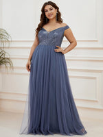A woman wearing an elegant off-shoulder slate blue gown with a sequined bodice stands confidently in a softly lit room, exuding grace and poise.
