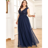 Woman in an elegant, sleeveless navy blue evening gown with a deep V-neckline stands in a bright, spacious room. The dress has a shimmering, fitted bodice and flowing skirt.