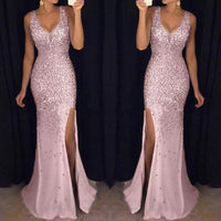 A woman in a shimmering, sleeveless pink gown with a deep v-neck and high slit stands in front of a dark background, conveying elegance and glamour.