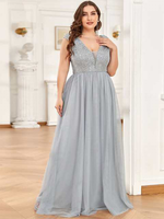 A woman in an elegant, sleeveless silver gown with a sequined bodice stands in a bright room with white walls and wooden floors, exuding a sophisticated and confident aura.