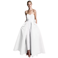A woman in a modern white gown stands confidently. The strapless dress has a flowing overskirt revealing fitted pants, combining elegance and contemporary style.