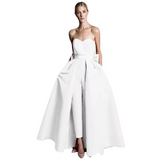 A woman in a modern white gown stands confidently. The strapless dress has a flowing overskirt revealing fitted pants, combining elegance and contemporary style.